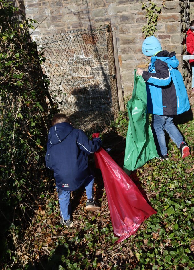 Kinder suchen mit einer grünen und einer roten Mülltüte im Beet an der Stadtmauer nach Müll.