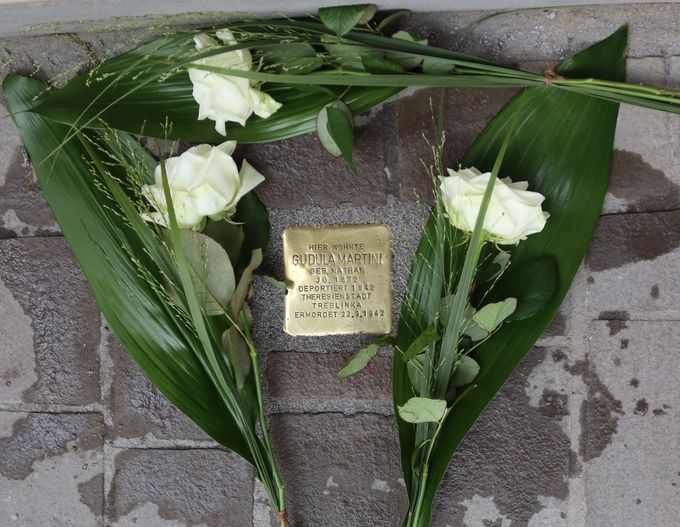 Ein Stolperstein aus Messing mit der Inschrift „Hier wohnte Gudula Martini“ liegt im Pflaster. Links und rechts davon liegen weiße Rosen mit grünen Blättern.