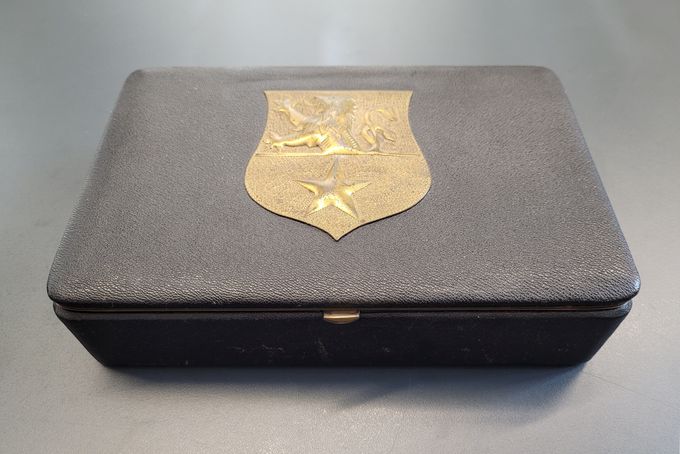 Ein alte, mit schwarzem Lederimitat überzogene Kiste mit dem goldfarbenen Wappen der Stadt Bad Münstereifel als Relief.