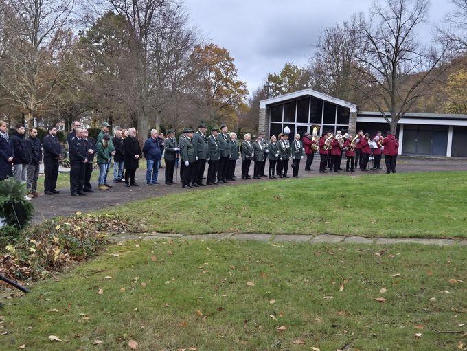 Schützen und Musikverein in Uniformen sowie weitere Personen stehen auf dem Weg neben einer Wiese auf dem Friedhof.