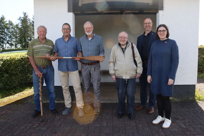 Sechs Personen stehen nebeneinander und schauen in die Kamera. Drei von ihnen halten ein hölzernes Paddel in den Händen.