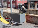 Die Gabel eines Baggers fährt einen Blumenkübel ins Bild. Ihr gegenüber steht ein Mann in Arbeitskleidung, hinter ihm ist eine hüfthohe Mauer. Er guckt auf den Blumenkübel und dirigiert den Baggerfahrer per Handzeichen.
