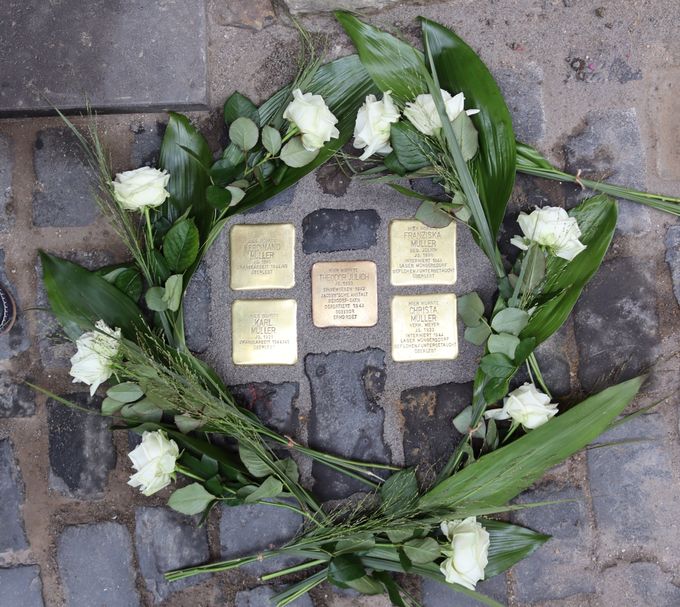 Fünf quadratische Stolpersteine aus Messing liegen im Kopfsteinpflaster, vier davon um einen mittleren Stein angeordnet. Rundherum liegen weiße Rosen und grüne Blätter.