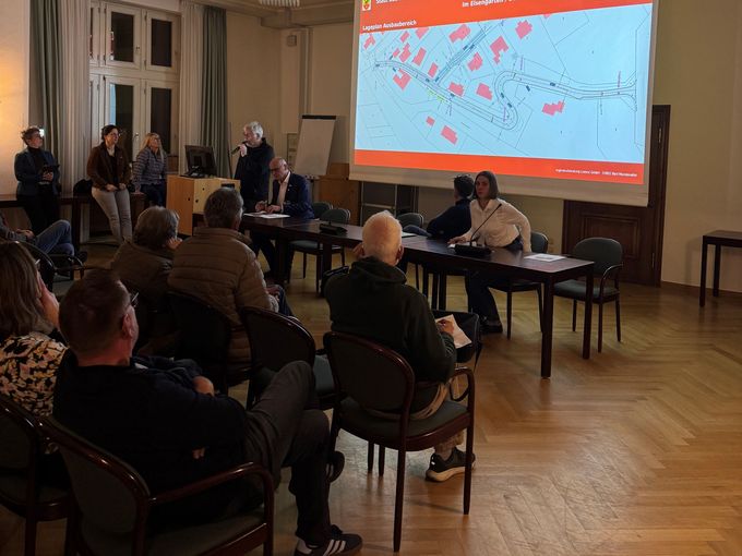 Im abgedunkelten Saal sitzen Menschen auf den Stühlen und schauen auf die Pläne, die auf einer Leinwand gezeigt werden.