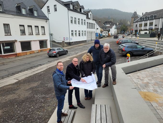 Fünf Personen stehen auf der Freitreppe am Bahnhof und schauen auf den Bauplan für die Kölner Straße. Im Hintergrund sieht man die Flutschäden an der Straße noch deutlich.