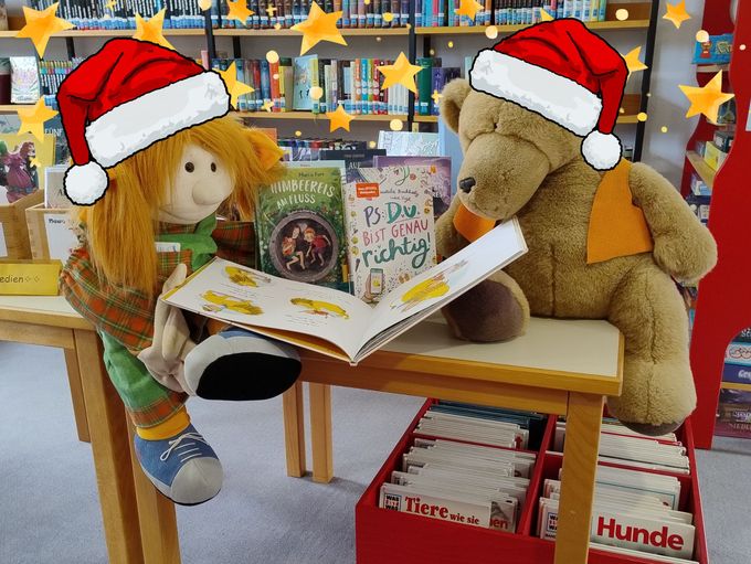 Eine Puppe (links) und eine Teddybär (rechts) sitzen in der Bücherei auf einem Tisch und schauen in ein Bilderbuch. Auf dem Tisch und im Regal im Hintergrund stehen Bücher. Das Foto wurde so bearbeitet, dass beide Figuren eine gezeichnete, rot-weiße Weihnachtsmannmütze tragen.