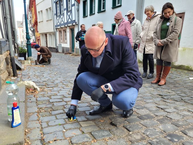 Bürgermeister Sebastian Glatzel und im Hintergrund Michael Meyer knien auf dem Pflaster und reinigen Stolpersteine. Daneben stehen Zuschauer.