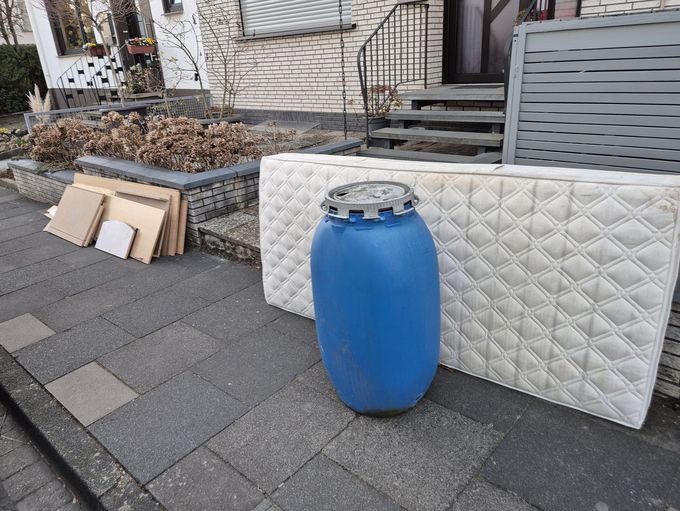 Auf einem Bürgersteig stehen eine Matratze, die an eine kniehohe Grundstücksmauer gelehnt ist, und ein blaues Plastikfass. Abseits davon stehen Teile eines Holzschranks.