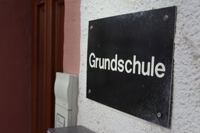 Ein simples braunes Schild mit der Aufschrift "Grundshchule" hängt an der weißen Außenwand des Schulgebäudes neben der hölzernen Eingangstür.