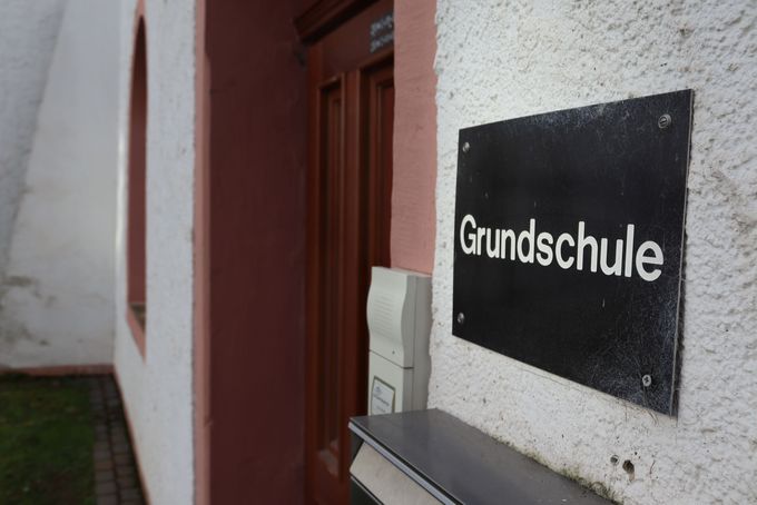 Ein simples braunes Schild mit der Aufschrift "Grundshchule" hängt an der weißen Außenwand des Schulgebäudes neben der hölzernen Eingangstür.