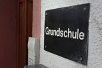 Ein simples braunes Schild mit der Aufschrift "Grundshchule" hängt an der weißen Außenwand des Schulgebäudes neben der hölzernen Eingangstür.