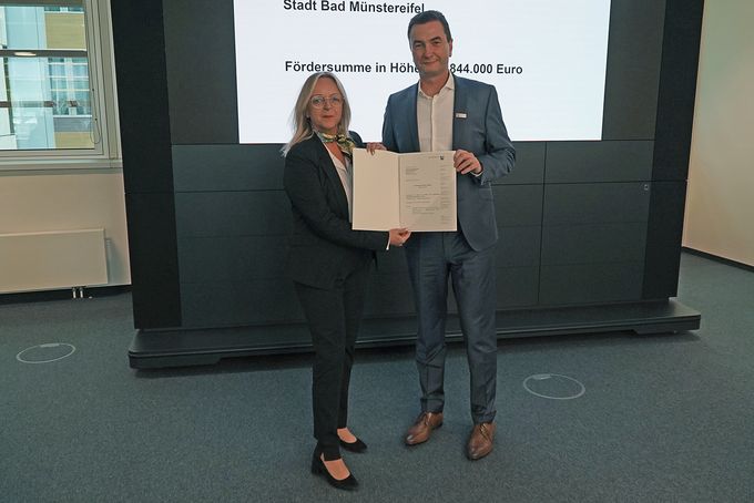 Regierungspräsident Dr. Thomas Wilk überreicht Bürgermeisterin Sabine Preiser-Marian den Förderbescheid in Form eines geöffneten DIN A4-Mappe.