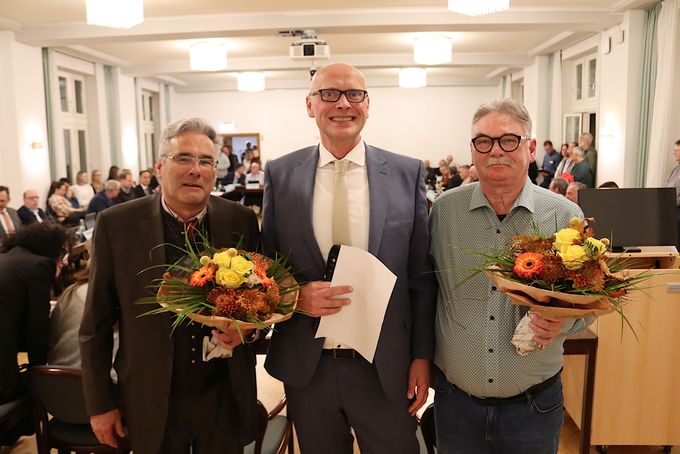 Ludger Müller, Bürgermeister Sebastian Glatzel und Rolf Winter stehen nebeneinander. Müller und Winter mit Blumensträußen.