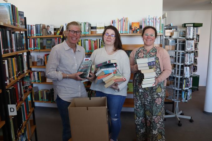 Drei Frauen stehen in einer Bücherei zwischen Bücherregalen und halten mehrere Bücher in den Händen. In der Mitte steht ein Karton mit weiteren Büchern, der für den Umzug gepackt wird. Im Hintergrund sind gefüllte Regale und ein Medienständer zu sehen.