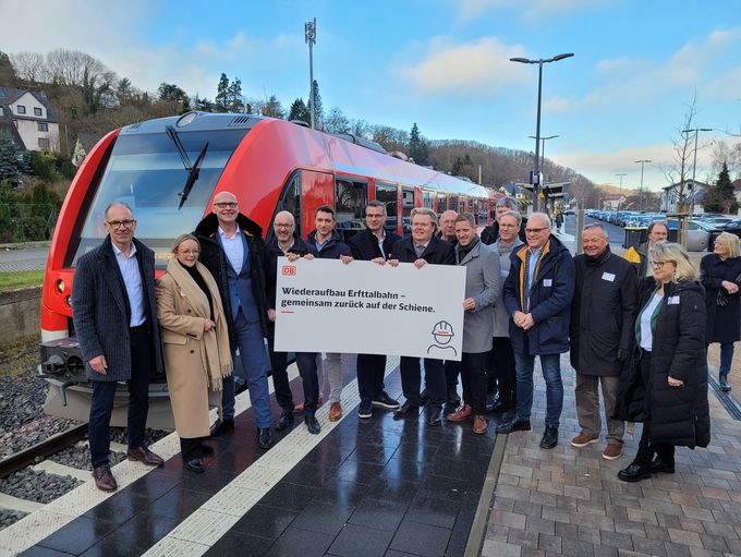 Viele Menschen stehen am Bahnsteig. Im Hintergrund der Zug, im Vordergrund ein großes Schild mit der Aufschrift: "Wiederaufbau Erfttalbahn - gemeinsam zurück auf der Schiene"