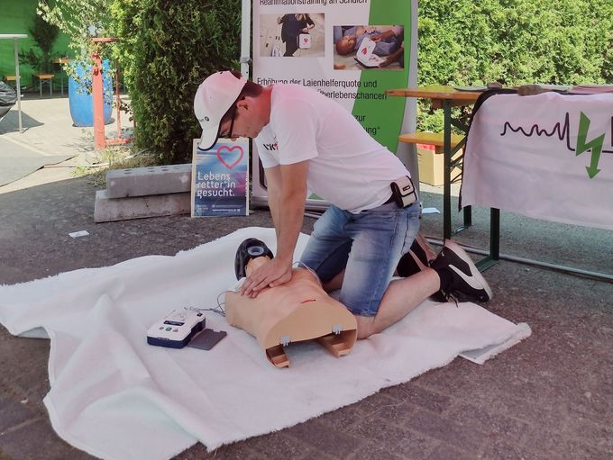 Fabian Brühl kniet draußen unter dem roten Dach eines entfaltbaren Pavillons auf dem gepflasterten Boden,unter ihm liegt eine weiße Decke. Er demonstriert die Wiederbelebung am Torso eines Puppe, daneben liegt der Defibrillator.