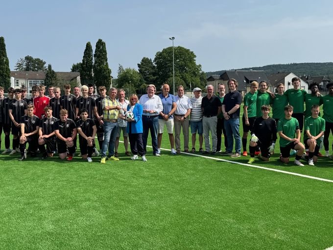Viele Menschen stehen in mehreren Reihen auf dem neuen Kunstrsenplatz in Arloff. Rechts und links sind einige Mitglieder von zwei Fußballmannschaften zu sehen. In der MItte sind mehrere Menschen, darunter Bürgermeisterin Sabine Preiser-Marian und Stefan Cremer,  Vorsitzender des RSV Arloff-Kirspenich. Preiser-Marian überreicht Cremer einen offiziellen Fußball zur EM 2024 und den Schlüssel zur Sportanlage.