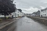 Blick auf eine regennasse Straße. Rechts und links Häuserzeilen. Rechts ist eine ca. 1,20 Meter hohe Mauer zu sehen.