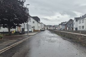 Blick auf eine regennasse Straße. Rechts und links Häuserzeilen. Rechts ist eine ca. 1,20 Meter hohe Mauer zu sehen.