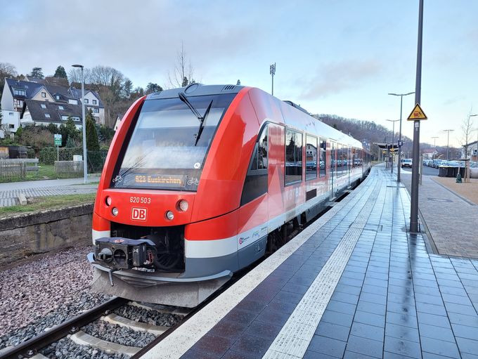 Ein roter Regionalzug der Deutschen Bahn steht am Bahnsteig im Bahnhof Bad Münstereifel. Der Zug zeigt als Ziel „Euskirchen“. Der Bahnsteig ist gepflastert und mit Leitstreifen für sehbehinderte Menschen ausgestattet, im Hintergrund sind Wohnhäuser und Bäume zu sehen.