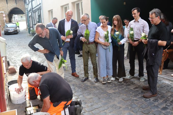 Auf einer Kopfsteinpflastergasse in der Altstadt stehen mehrere Menschen mit weißen Rosen in den Händen und blicken nach unten. Links knien zwei Männer in orangefarbener Warnkleidung am Boden und verlegen die Stolpersteine im Pflaster..