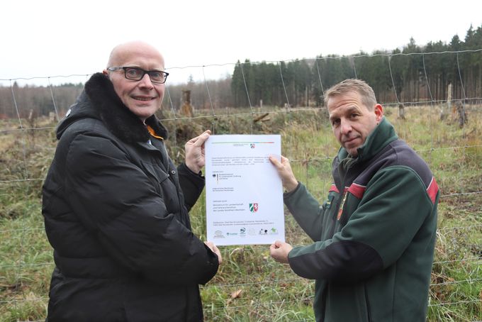 Bürgermeister Sebastian Glatzel und Forstbüroleiter Stefan Lott halten ein Plakat an einen Zaun. Im Hintergrund ist eine große Lichtung im Wald.