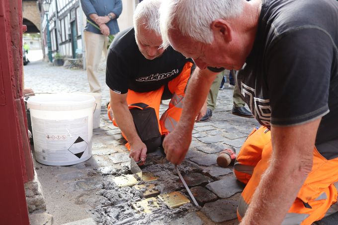 Zwei Bauhof-Mitarbeiter knien auf dem Kopfsteinpflaster und setzen mit Kelle und Spachtel mehrere Messingplatten in Mörtel ein. Neben ihnen steht ein weißer Eimer.