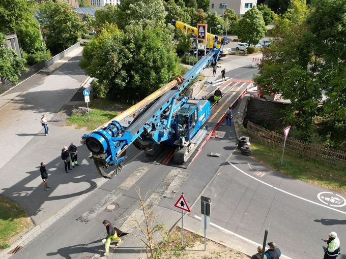 Luftbild eines sehr großen, blauen Baufahrzeugs, das über die Erftbrücke am Sittardweg fährt. Unter dem Fahrzeug ist der vorübergehende Überbau der Brücke zu erkennen.