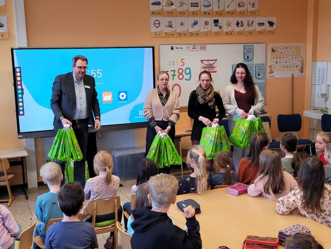 Andreas Bouß von Volksbank Euskirchen, Bürgermeisterin Sabine Preiser-Marian, Annika Simon und Sonja Aufdermauer von der Stadtbücherei haben viele grüne ABC-Tüten aus Papier in den Händen, ums sie an die Erstklässler zu verteilen, die im Klassenraum vor ihnen sitzen.