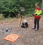 Ein Mann in Warnjacke steuert eien Drohne knapp über dem Waldboden. Daneben ein Monitor auf einem Stativ.