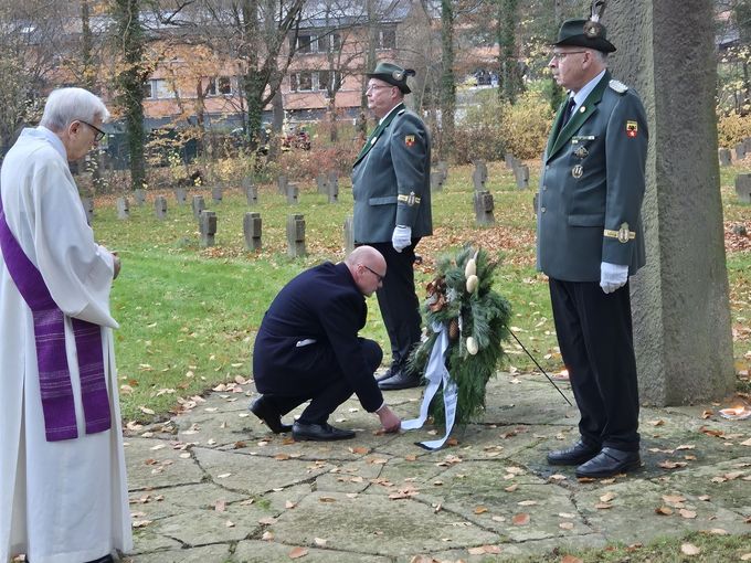 Bürgermeister Sebastian Glatzel hockt vor dem Ehrenmal auf dem Boden und zieht die Schleife des Kranzes zurecht. Zwei Schützen stehen neben dem Ehrenmal. Dabei steht ein katholischer Diakon.
