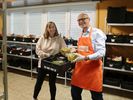 Gabi Schönemann hält Bürgermeister Sebastian Glatzel einen Plastikkorb mit Lebensmitteln in. Der Bürgermeister nimmt eine Packung Weintrauben heraus.