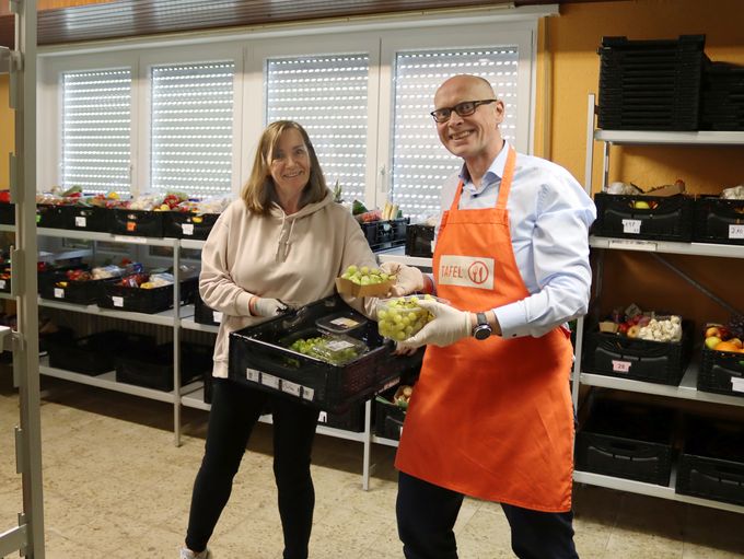Gabi Schönemann hält Bürgermeister Sebastian Glatzel einen Plastikkorb mit Lebensmitteln in. Der Bürgermeister nimmt eine Packung Weintrauben heraus.