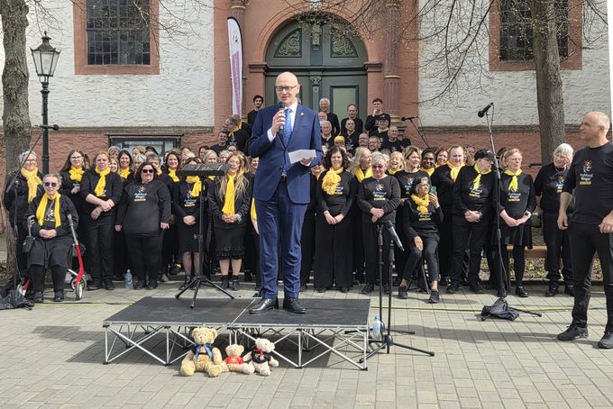 Bürgermeister Sebastian Glatzel im blauen Anzug steht auf einer kleinen Bühne vor einer Kirche und spricht in ein Mikrofon. Hinter ihm steht ein großer Chor mit vielen Sängerinnen und Sängern in schwarzer Kleidung und gelben Schals. Mikrofone und Notenständer sind aufgebaut, im Vordergrund stehen drei Stofftiere vor der Bühne.