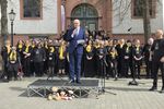 Bürgermeister Sebastian Glatzel im blauen Anzug steht auf einer kleinen Bühne vor einer Kirche und spricht in ein Mikrofon. Hinter ihm steht ein großer Chor mit vielen Sängerinnen und Sängern in schwarzer Kleidung und gelben Schals. Mikrofone und Notenständer sind aufgebaut, im Vordergrund stehen drei Stofftiere vor der Bühne.