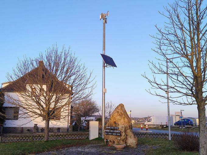 Auf einem kleinen Platz am Straßenrand steht ein hoher Sirenenmast mit Lautsprechern und einem Solarpanel. Daneben befindet sich ein Gedenkstein, im Hintergrund sind ein Wohnhaus, Bäume und eine Straße mit Fahrzeugen zu sehen.