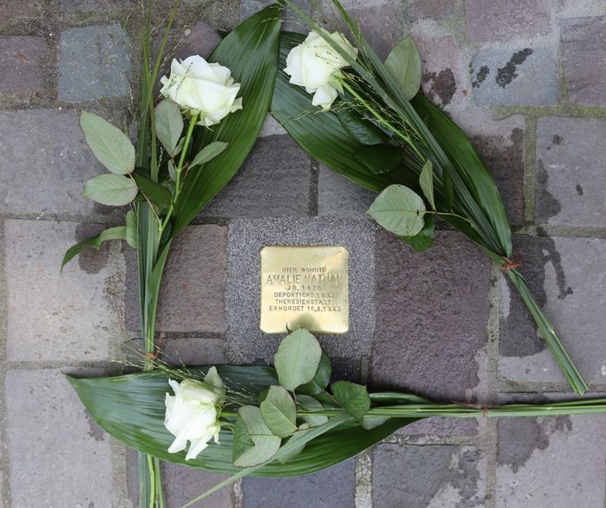 Ein Stolperstein aus Messing mit der Inschrift „Hier wohnte Amalie Nathan“ liegt im Pflaster. Links und rechts davon liegen weiße Rosen mit grünen Blättern.