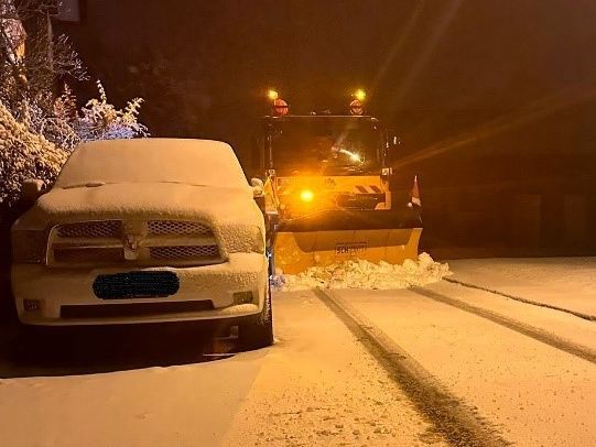 Ein Räumfahrzeug des Winterdienstes fährt nachts auf einer schneebedeckten Straße an einem geparkten Auto vorbei.