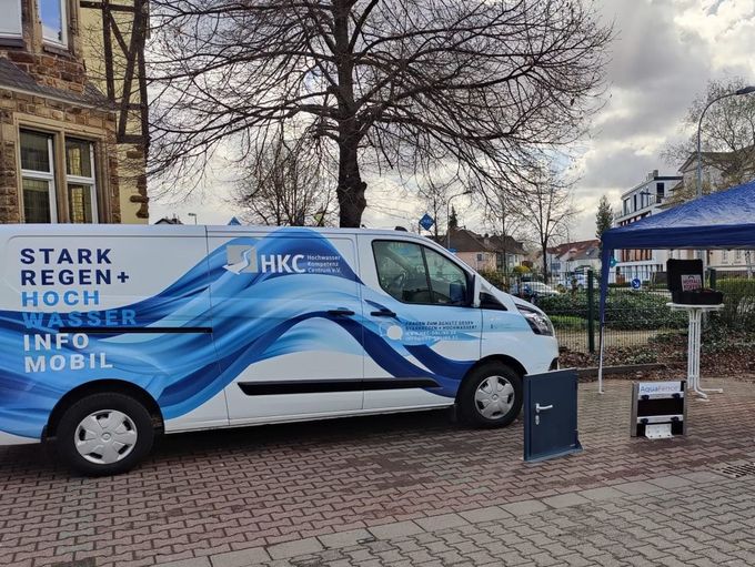Ein Transporter, bedruckt mit blauen stilisierten Wellen, steht in einer unbekannten Stadt neben einem blauen Pavillondach.