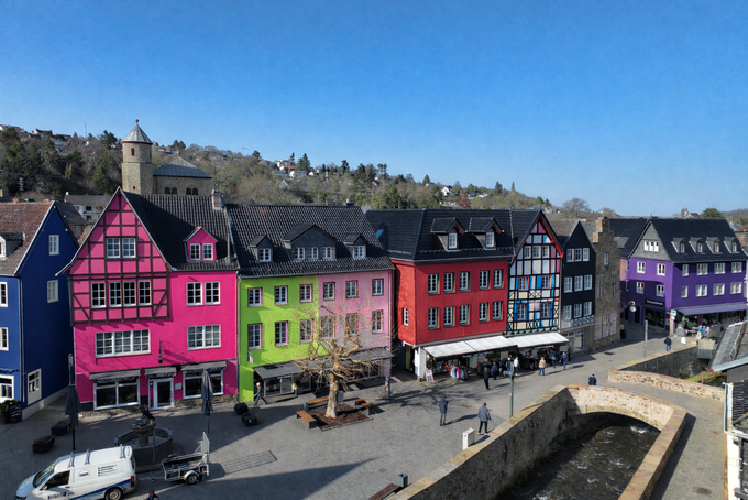 Blick auf den Markt in Bad Münstereifel mit einer Reihe eng stehender Häuser, die in auffälligen Farben wie Pink, Grün, Gelb, Rot und Lila gestrichen sind. Die Farben wurden digital bearbeitet. Vor den Gebäuden verläuft ein Platz mit wenigen Passanten, rechts fließt die Erft entlang einer steinernen Uferbefestigung.